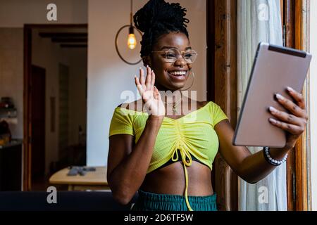 Allegra giovane femmina nera in cima raccolto e occhiali ondeggianti mani e sorridi mentre si ha una chat video sul tablet a. casa Foto Stock