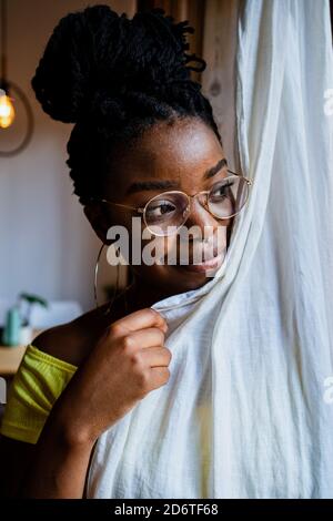 Giovane donna afro-americana pensiva in occhiali alla moda che coprono metà del suo corpo con tenda e guardando la macchina fotografica a casa Foto Stock