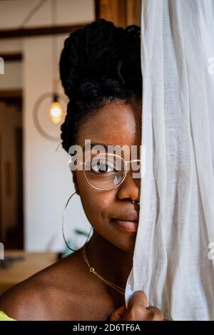 Giovane donna afro-americana pensiva in occhiali alla moda coprendo metà del viso con tenda e guardando la macchina fotografica a casa Foto Stock