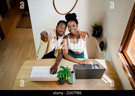 Dall'alto di positivo giovane afro-americano uomo e donna in abiti casual puntando la fotocamera e sorridendo mentre lavorano insieme su computer portatili a casa Foto Stock