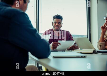 Uomo d'affari che discute lavoro di carta che ascolta il capo seduto dentro sala conferenze durante il team meting Foto Stock