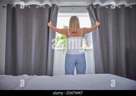 Vista posteriore della donna che indossa le tende di apertura Pajamas e l'aspetto Fuori dalla finestra di mattina Foto Stock