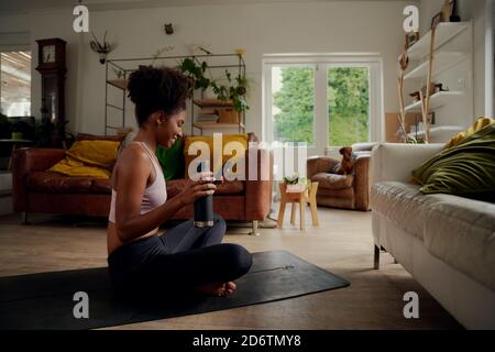 Sorridente giovane donna africana che tiene la bottiglia e l'uso dello smartphone durante allenati mentre sei seduto sul tappetino fitness a casa Foto Stock