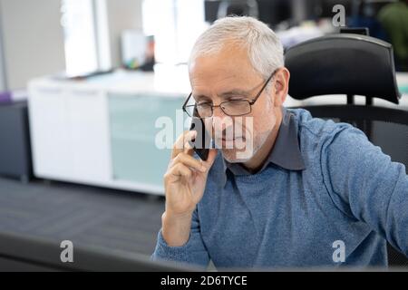Uomo d'affari senior che parla con lo smartphone mentre si siede sulla scrivania in un ufficio moderno Foto Stock