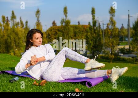 Bella donna bruna sportiva giaceva sul tappetino a verde soleggiato giorno e fare fitness abs crunches con palme chiuse mani Foto Stock