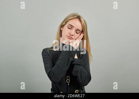 Dormire con la testa appoggiata su mani aggrappate come sogni dolci, occhi chiusi, primo piano, gesturante segno di addormentamento, riposo. Giovane donna attraente, vestito maglione nero con occhi verdi, capelli biondi, sfondo Foto Stock