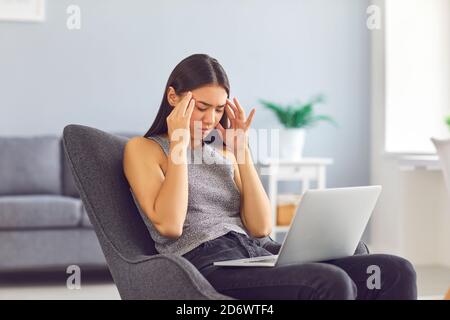 Stanca giovane donna strofinare templi che soffrono di mal di testa seduta dentro poltrona con computer portatile a casa Foto Stock