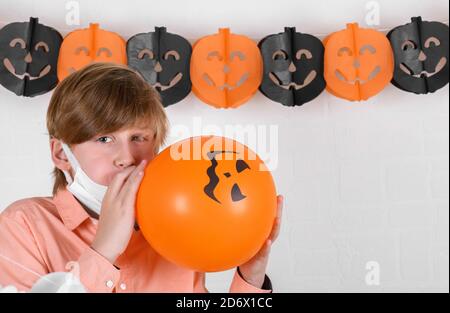 Un ragazzo biondo in maschera medica protettiva gonfia un pallone arancione, Happy Halloween sulla quarantena coronavirus pandemia Foto Stock
