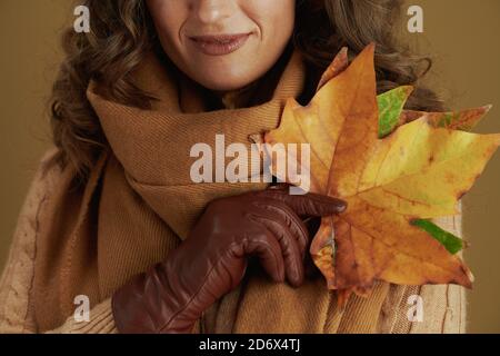 Ciao settembre. Primo piano su donna in sciarpa con guanti in pelle e foglie d'acero giallo autunnale su fondo in bronzo. Foto Stock