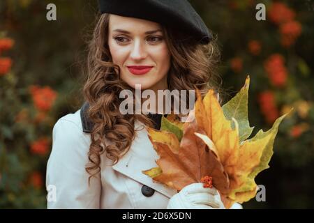 Ciao settembre. Elegante donna di mezza età in trench beige con foglie gialle autunnali all'aperto in città in autunno. Foto Stock