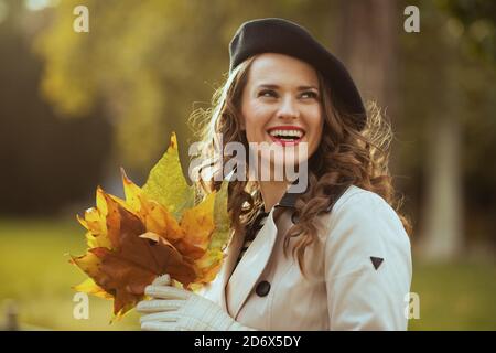 Ciao settembre. Felice donna di stile in trincee camice beige e beretto nero con foglie di autunno giallo all'aperto sulla strada della città in autunno. Foto Stock