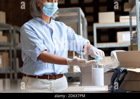 Donna matura in magazzino che indossa una maschera con igienizzatore al lavoro. Foto Stock