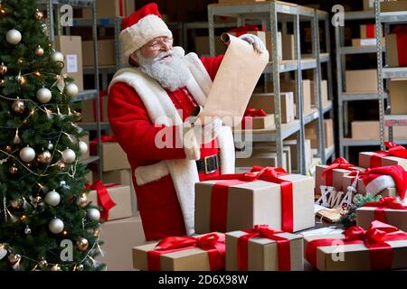 Felice Babbo Natale che indossa costume che legge lista dei desideri in piedi in officina. Foto Stock