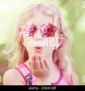 Giovane ragazza che posa in un elegante pentagonale rosa a forma di occhiali da sole all'aperto. Carino adorabile bambino caucasico con lunghi capelli biondi che inviano bacio d'aria. Co Foto Stock