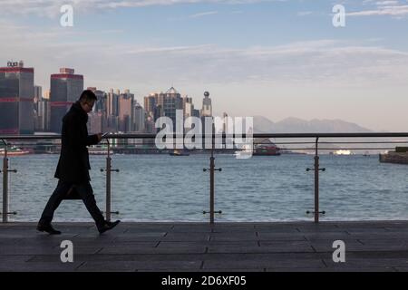 Un uomo d'affari, utilizzando uno smartphone, cammina lungo il lungomare della baia di Hong Kong a Tsim Sha Tsui, Hong Kong, Cina. Foto Stock