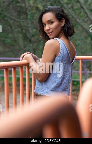 Bella giovane Latina con capelli corti ondulati, in piedi appoggiati a una ringhiera, mostrandola dietro, indossando una blusa blu, pantaloni neri, stile casual Foto Stock