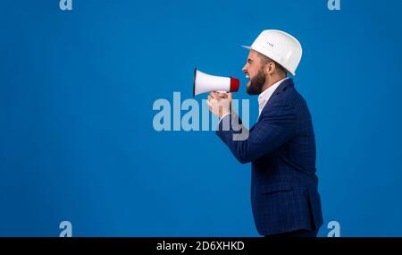 un operaio di costruzione di mezza età in una tuta e un casco da costruzione parla in un megafono su uno sfondo blu isolato . un giovane Foto Stock