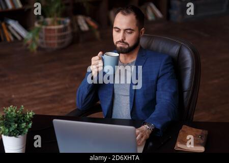 Caffè fresco al lavoro. Giovane sicuro in abbigliamento casual elegante che lavora su un computer portatile e beve una bevanda calda mentre si siede al lavoro Foto Stock