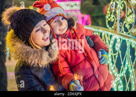 Piccolo ragazzo e sua madre con sparklers vicino all'abeto gigante e illuminazione di Natale sul mercato di Natale. Vacanze di Natale in fiera Foto Stock