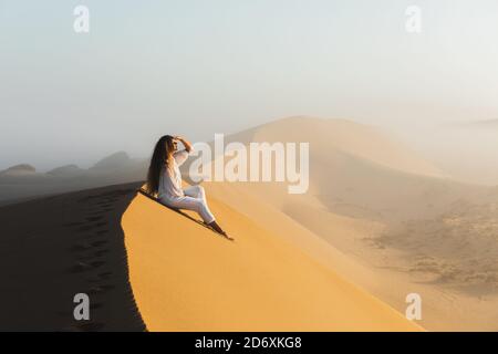 Destinazione da sogno, viaggio e wanderlust concetto. Donna felice ispirata dalla fantastica alba nelle dune di sabbia del deserto del Sahara, Marocco. Nebbia al mattino e luce calda. Foto Stock