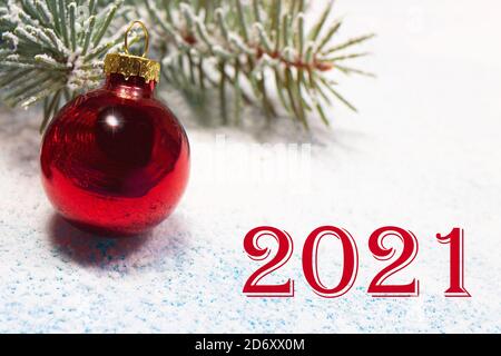 Bella carta di Capodanno con l'iscrizione dei numeri 2021, una palla rossa e un ramo innevato di abete rosso nella neve. Foto Stock