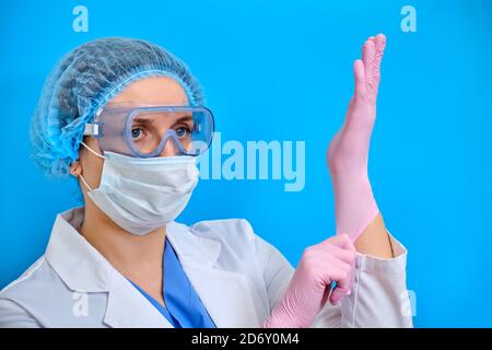 Infermiere in occhiali indossa guanti protettivi per il lavoro, sfondo blu. Un medico in un camice bianco e attrezzature protettive per il trattamento dei pazienti Foto Stock