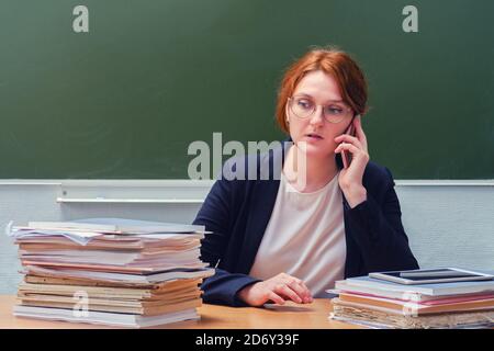 L'insegnante si siede a una scrivania e parla al telefono la lavagna della scuola Foto Stock