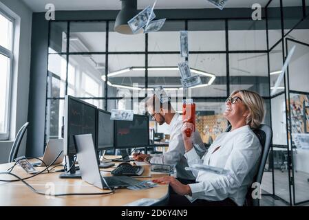 Gettando denaro in aria con un dispositivo speciale. Due agenti di borsa in abiti formali hanno una pausa e divertimento in ufficio Foto Stock