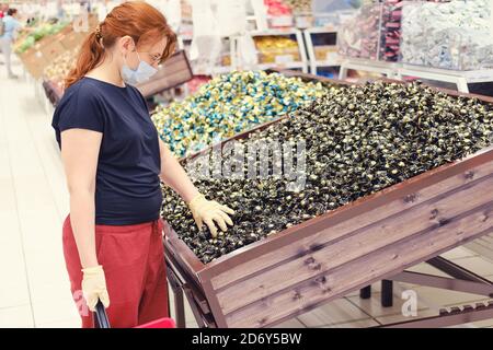 Una donna in una maschera medica sceglie i dolci nel negozio. Acquistare caramelle in un supermercato. Visitare luoghi pubblici durante la pandemia del coronavirus Foto Stock