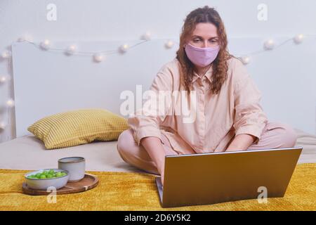 Donna in maschera medica è seduta con il computer portatile sul letto di casa, la prima colazione del mattino. Ragazza lavora al computer online durante l'isolamento a causa di coronavirus, co Foto Stock