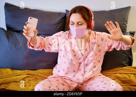 Una donna ascolta musica e balli mentre si siede sul letto durante una malattia con il virus dell'influenza covid-19. Ragazza in pigiama, cuffie e un protettori Foto Stock