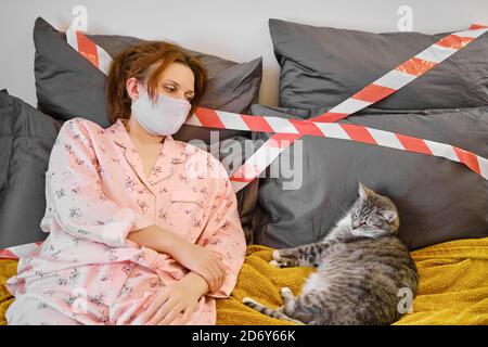 Una donna in una maschera medica casalinga si trova con un gatto durante una malattia del coronavirus, concetto. Ragazza dai capelli rossi in pigiama su un letto con un nastro di arresto durante Foto Stock