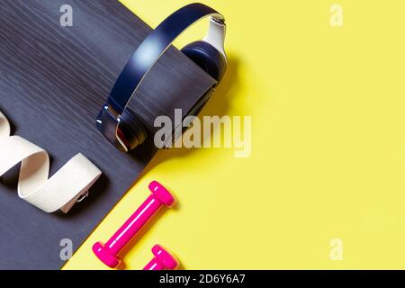 Flat Lay attrezzature sportive e fitness, manubri rosa, cuffie, tappetino nero per yoga e cinturino yoga su sfondo giallo. Vista dall'alto con spazio per t Foto Stock
