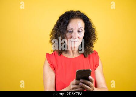 Donna di mezza età che indossa una camicia casual che si erge su giallo isolato sms di sottofondo con il suo telefono Foto Stock