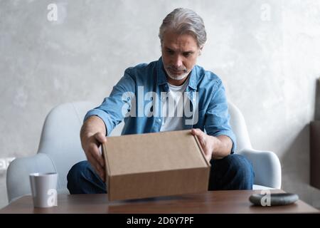 Uomo sorridente consumatore aperto scatola di cartone Get pacco postale, cliente maschile ricevere confezione di cartone sedersi sul divano a casa Foto Stock