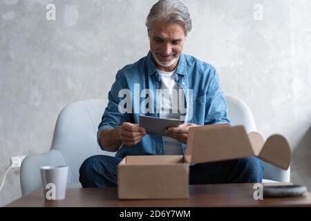 Uomo sorridente consumatore aperto scatola di cartone Get pacco postale, cliente maschile ricevere confezione di cartone sedersi sul divano a casa Foto Stock