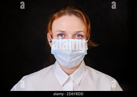 Una donna con occhi rossi in una camicia bianca e una maschera medica su sfondo nero. Persone catturate in isolamento a causa del virus dell'influenza Foto Stock