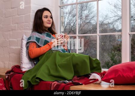 Una ragazza si siede su un davanzale vicino ad una grande finestra in inverno. Albero di Natale e regali di Capodanno 2021 Foto Stock