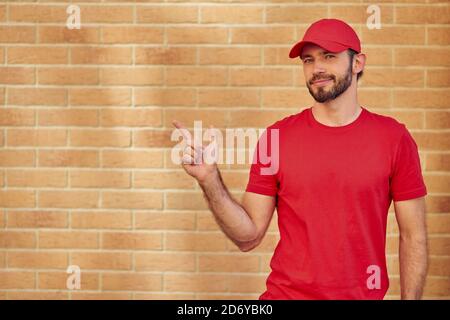 Giovane allegro caucasico maschio ciurier in uniforme rossa facendo gesto con le dita su muro di mattoni sfondo, copia spazio. Concetto di consegna degli alimenti Foto Stock