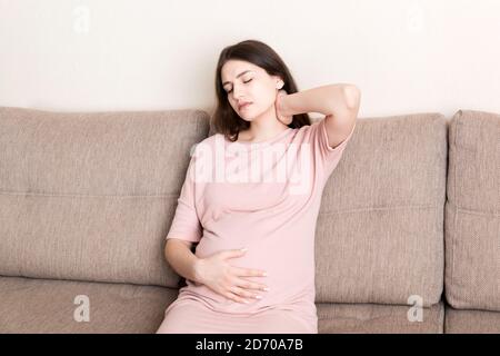 Giovane donna incinta che soffre di collo sul divano a casa. Concetto di assistenza sanitaria per la maternità. Foto Stock
