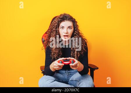 Donna seduta dietro la sedia da gioco nelle mani che tiene in rosso gamepad su sfondo giallo Foto Stock