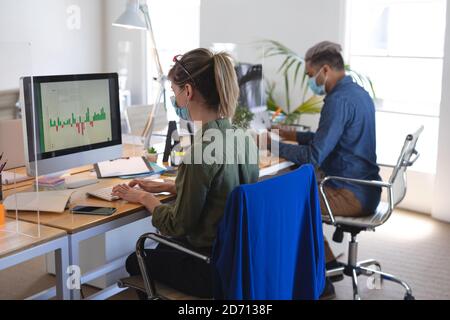 Donna caucasica seduta alla scrivania in un ufficio moderno con la sua collega, indossando una maschera facciale e utilizzando un computer. Salute e igiene sul luogo di lavoro d Foto Stock