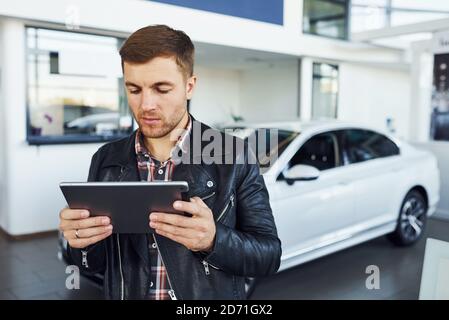Uomo in giacca nera si trova all'interno del salone auto con tablet nelle mani Foto Stock