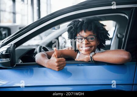 Mostra il pollice verso l'alto. La giovane donna afroamericana si siede all'interno della nuova vettura moderna Foto Stock