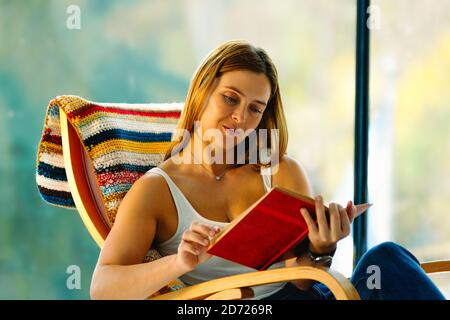 Giovane donna attraente legge un libro, dondolando in una sedia dondolo comoda. Un buon passatempo migliora il tuo umore. Foto Stock