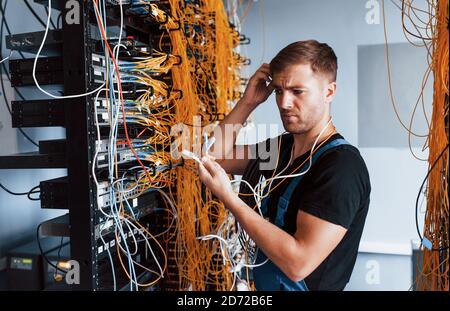 Giovane uomo in uniforme si sente confuso e alla ricerca di un soluzione con apparecchiature internet e cavi nella sala server Foto Stock