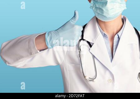 Medico femminile in maschera protettiva, camice bianco, stetoscopio e guanti di gomma che mostrano il pollice in su Foto Stock