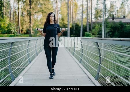 Donna europea sportiva in camicia nera t e leggings, cammina attraverso il ponte, tiene due manubri, lavori su bicipiti di addestramento, treni all'aperto, ha pensefu Foto Stock