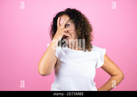 Donna di mezza età che indossa una camicia bianca casual che si erge su uno sfondo rosa isolato sbirciando in shock coprendo viso e occhi con la mano, guardando attraverso Foto Stock