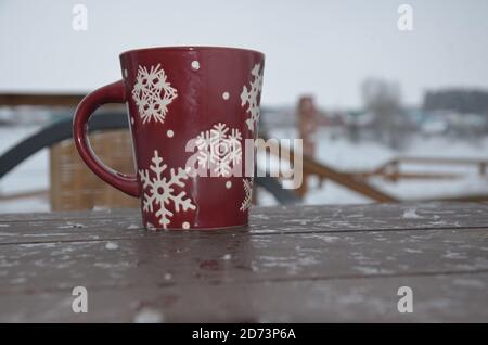 Coppa rossa o borgogna con un modello di fiocchi di neve sullo sfondo di una foresta invernale. Nevicare. Capodanno. Natale. Tazza con una bevanda calda al vento Foto Stock